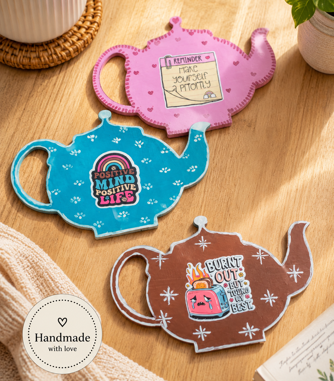 Three colorful teapot-shaped coasters on a wooden surface with a cup and plant in the background.