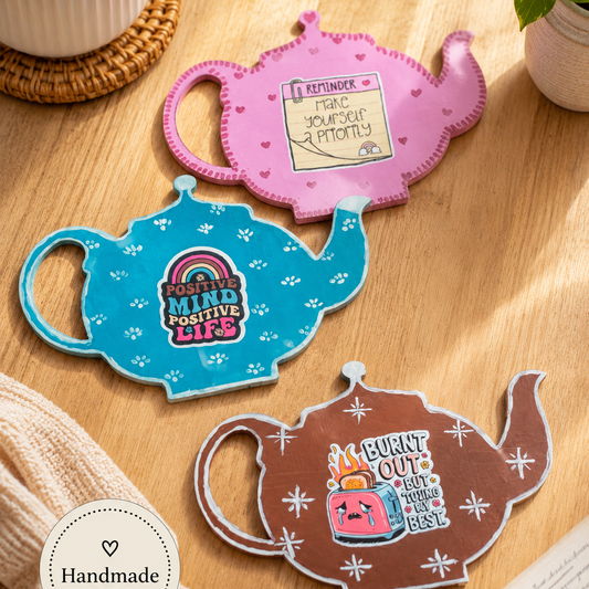 Three colorful teapot-shaped coasters on a wooden surface with a cup and plant in the background.